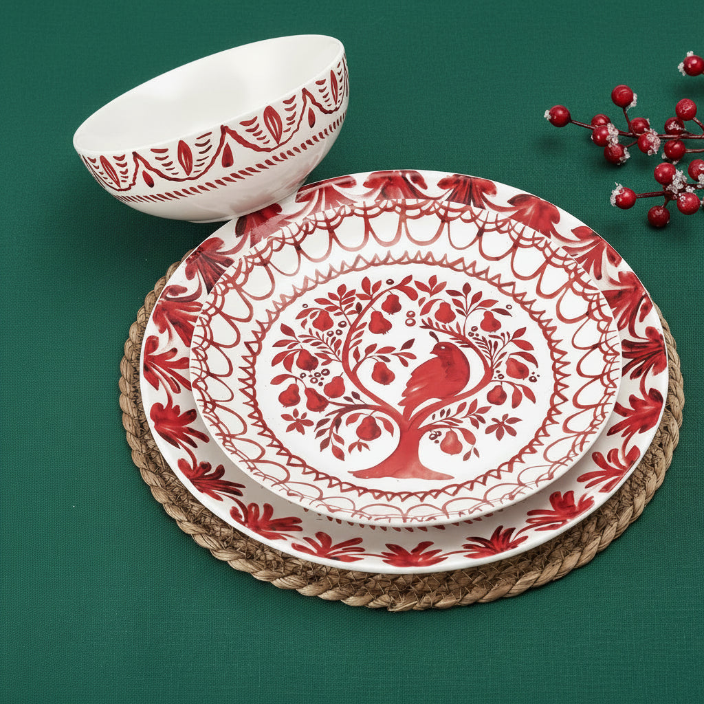 A red and white patterned dinnerware set on a light wood table, featuring a large plate, a matching bowl, a woven placemat, a rolled red napkin, and red berries as styling props.