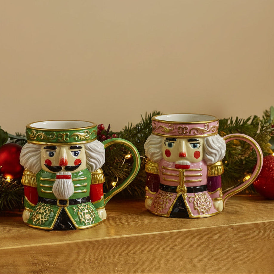 Two ceramic nutcracker mugs with colorful painted faces and ornate handles, displayed on a wooden surface with holiday greenery and red ornaments in the background.