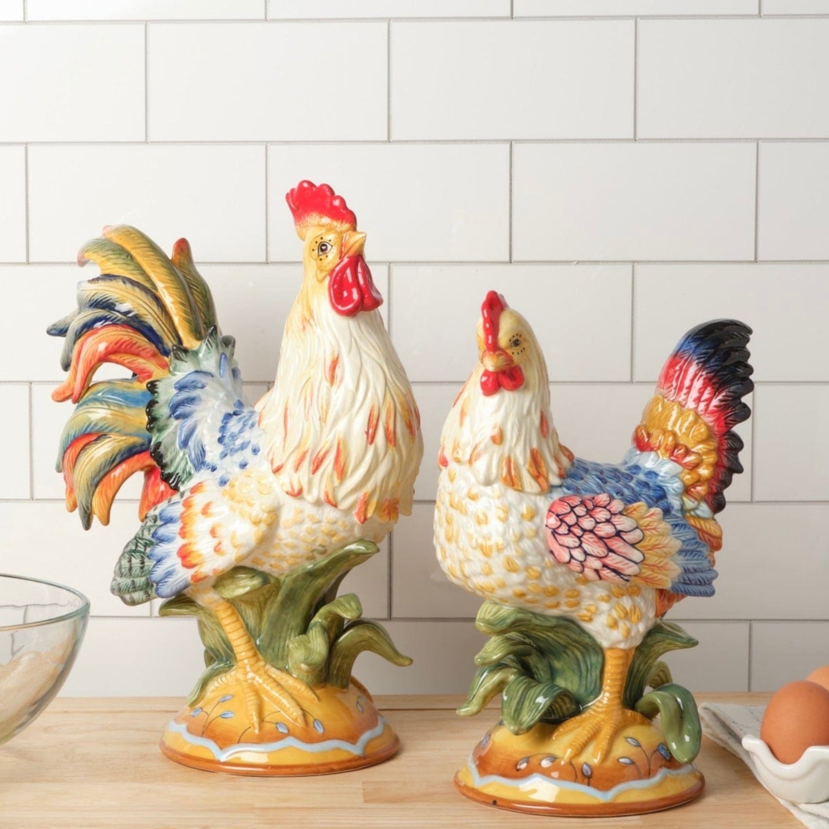 Two colorful ceramic rooster figurines on a kitchen counter with eggs and a whisk.