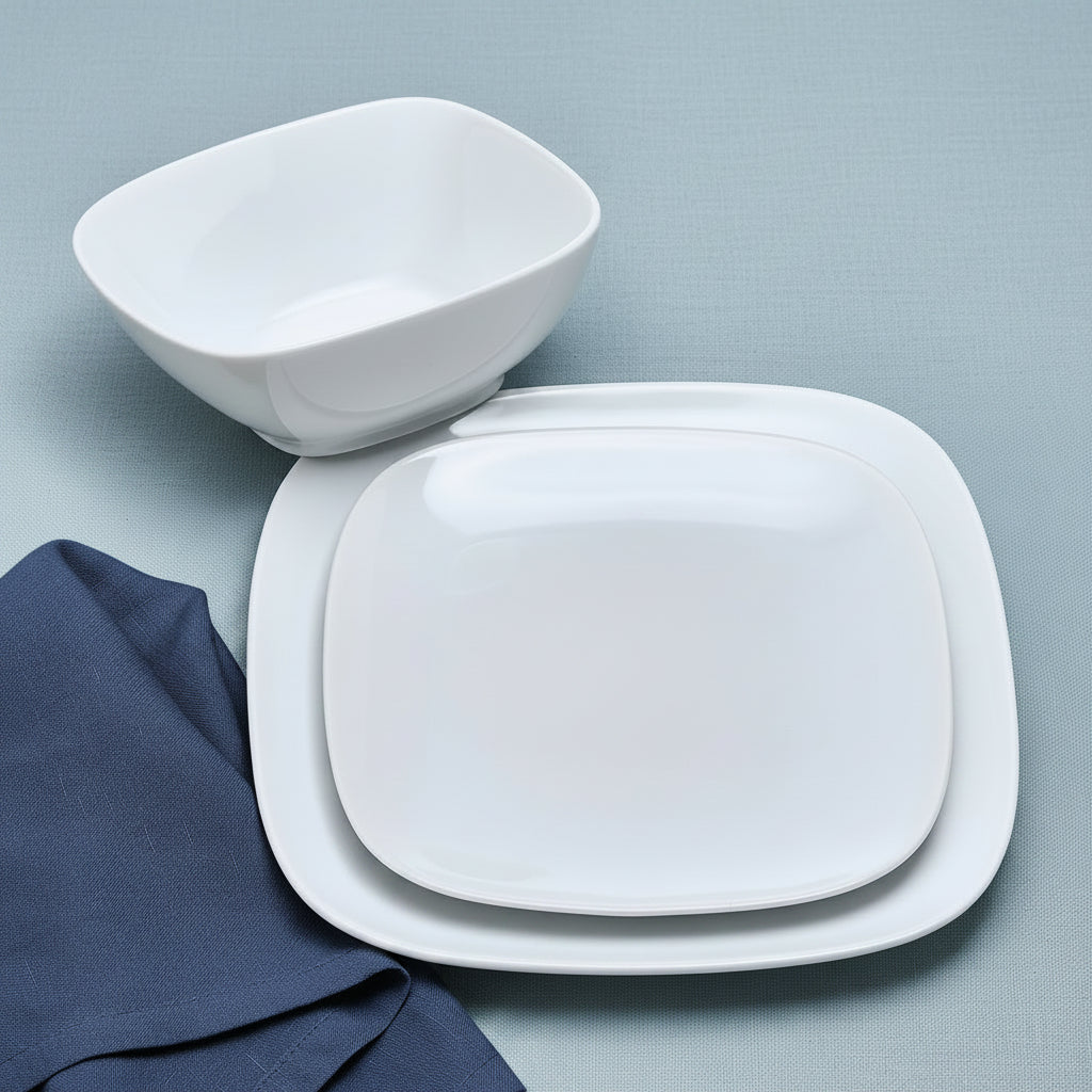 A set of white ceramic square plates with rounded corners, including a shallow bowl and two stacked serving plates, arranged on a wooden tabletop with a green napkin and white flowers.